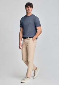 Herenoutfit met een aansluitend marineblauw t-shirt, beige broeken en lichtgekleurde sneakers. Het t-shirt heeft een klein logo op de borst.