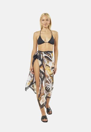 Donna che indossa un top bikini nero, un sarong con motivo astratto legato in vita e sandali neri, in piedi davanti a uno sfondo semplice.