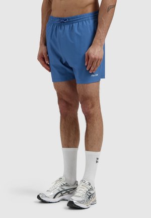 Mannelijk model dat blauwe sportshorts draagt met het logo "Pure Polis", witte sokken en zilveren hardloopschoenen, staand tegen een effen achtergrond.