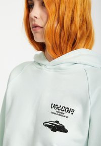 Ljusmyntgrön hoodie med en framficka i känguru-stil, snörning i huvan och svart grafisk tryck med en UFO och text på vänster bröst.