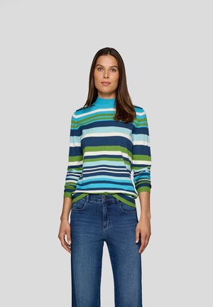 Frau mit langen Haaren, die einen gestreiften Pullover in Blau, Grün und Weiß sowie eine blaue Jeans trägt, steht vor einem schlichten grauen Hintergrund.