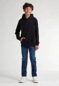 Černá mikina s kapucí a klokaní kapsou, nošená s modrými džínami a bílými teniskami. Tkanina je měkká a střih pohodlný.