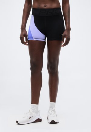 Αθλητής που φοράει μαύρο και μωβ σορτς συμπίεσης Nike Pro και λευκά αθλητικά παπούτσια Nike με λευκές κάλτσες, στέκεται μπροστά σε απλό φόντο.