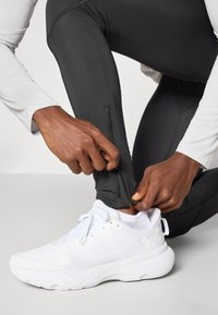 Scarpe da ginnastica bianche con tomaia in rete testurizzata e suola solida. Leggings neri aderenti con cerniere alla caviglia. Mani che regolano le cerniere.