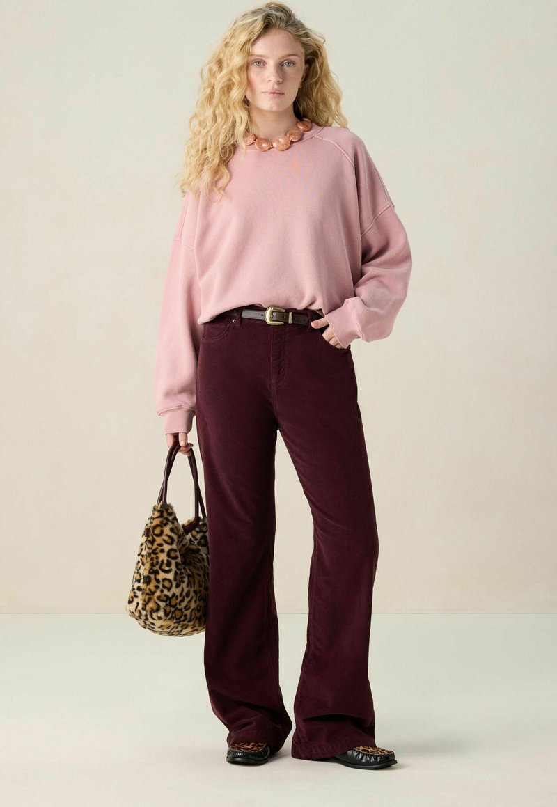 Femme aux cheveux blonds bouclés portant un sweat-shirt rose, un pantalon large bordeaux, des chaussures à imprimé léopard, un collier et tenant un sac à main à imprimé léopard.