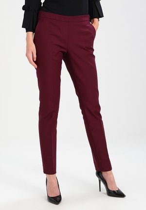 Mujer con pantalones ajustados burdeos con bolsillos y zapatos de tacón negros, combinados con una blusa negra con mangas con volantes.