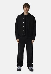 Giacca di jeans nera con bottoni argentati, indossata sopra una maglietta a maniche lunghe nera e abbinata a jeans neri a gamba larga e sneakers grigie.