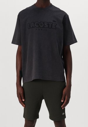 Dunkelgraues Lacoste T-Shirt mit schwarzem Logo, getragen mit dunklen Shorts mit kleinem Krokodil-Logo am linken Bein.