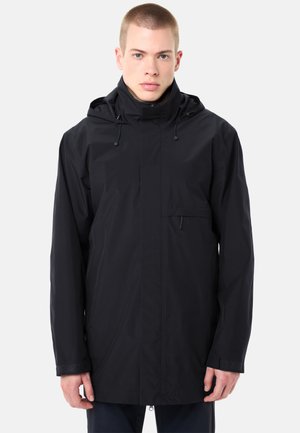 Jeune homme portant une veste imperméable noire à capuche avec une fermeture éclair frontale et des fermetures auto-agrippantes aux poignets, se tenant devant un fond blanc.