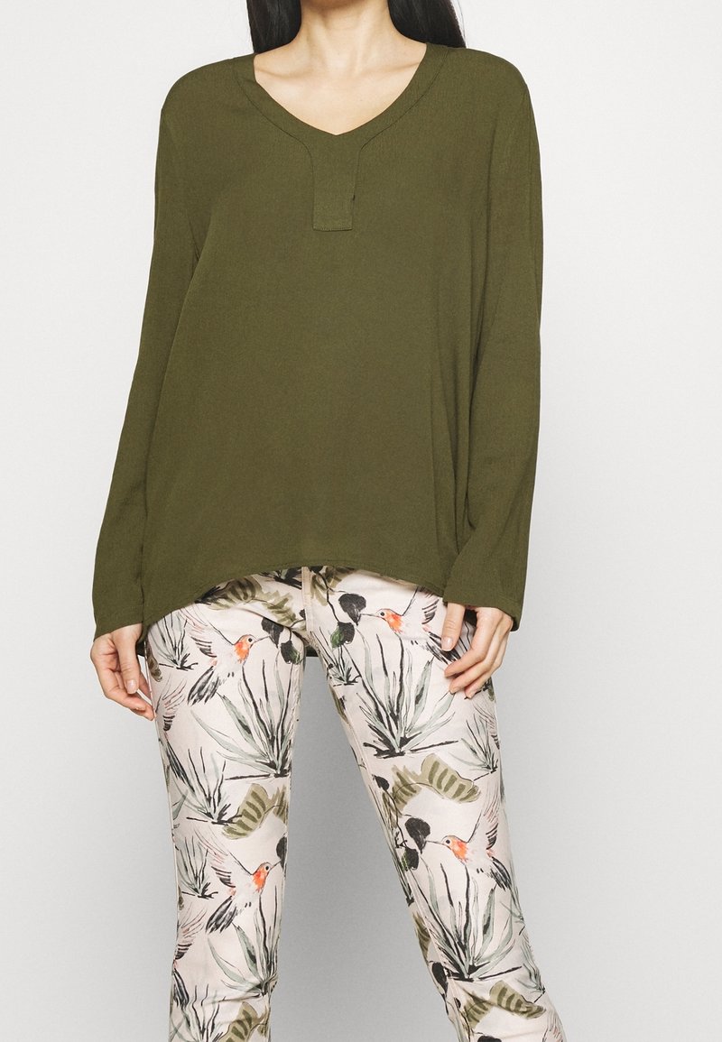 Femme portant un haut à manches longues vert olive avec un décolleté en V échancré et un pantalon crème orné d'un motif de colibri vert et orange.