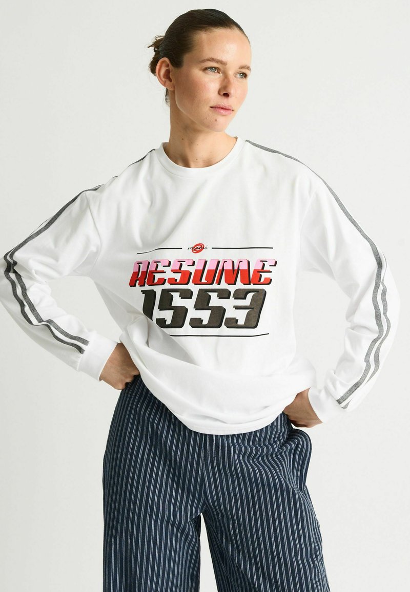 Hölgy hátrafogott hajjal, fehér hosszú ujjú ingben, amelyen a "RESUME 1553" felirat olvasható, és csíkos tengerészkék nadrágban, kezét csípőjén tartva pózol.