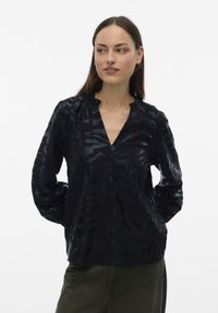 Vrouw met lang bruin haar, gekleed in een zwarte blouse met patronen en lange mouwen en een donkere broek, staand met de handen achter de rug, zijwaarts kijkend.