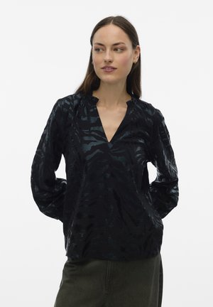 Vrouw met lang bruin haar, gekleed in een zwarte blouse met patronen en lange mouwen en een donkere broek, staand met de handen achter de rug, zijwaarts kijkend.