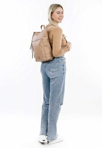 Zaino in pelle beige chiaro con chiusura a zip, tasca frontale piatta, spallacci imbottiti, forma rettangolare e superficie liscia, indossato da una persona.