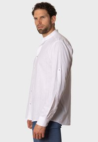Camisa blanca de manga larga hecha de tejido liviano, con cuello alto, cierre frontal de botones y detalles con botones en las mangas.