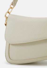 Ljust beige texturerat läderhandtag med en kurvad design, som har en klaffstängning och en guldtonad kedjestraps för bärande.