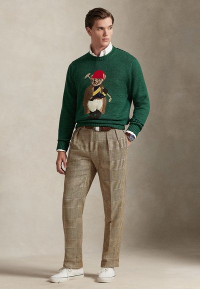 Πράσινο πλεκτό πουλόβερ με κεντημένο χαρακτήρα αρκούδας, συνδυασμένο με καφέ καρό παντελόνι και λευκά αθλητικά παπούτσια.