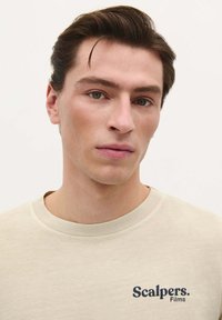 Beige Baumwoll-T-Shirt mit Rundhalsausschnitt und kurzen Ärmeln. Verfügt über ein navyblaues, gesticktes "Scalpers. Films"-Logo auf der Brust.