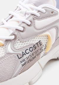 Κοντινή λήψη ενός λευκού και μπεζ αθλητικού παπουτσιού Lacoste με δίχτυ, λεπτομέρειες από σουέν τ και λευκά κορδόνια, με τυπωμένες συντεταγμένες στην πλαϊνή όψη.