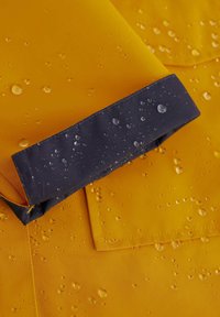 Geltona vandeniui atspari medžiaga su tamsiai mėlynu juostele, ant paviršiaus matomi vandens lašai, akcentuojantys tekstūrą ir medžiagos kokybę.