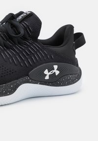 Sort atletisk sko med en tekstureret overdel, der har vertikale striber, en polstret krave og en hvidspraglet sål. Under Armour-logoet er synligt.