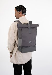 Rucsac gri cu capac rulant, fabricat dintr-un material durabil. Dispune de o cataramă frontală, buzunare laterale și un patch cu logo în partea de jos. Bretele ajustabile.