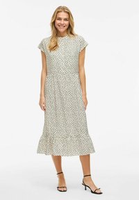 Prickig klänning i kräm och svart, med kort ärm, en figurnära bodice och en utsvängd nederdel. Tillverkad av lättviktsmaterial.