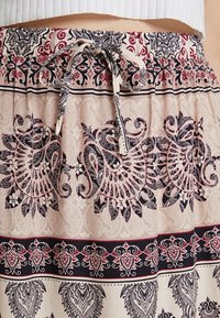 Hame trouseri elastik di dalam warna beige muda dengan pola geometri dan floral berwarna hitam dan burgundi yang rumit, dilengkapi dengan tali serut untuk pengaturan ukuran yang dapat disesuaikan.