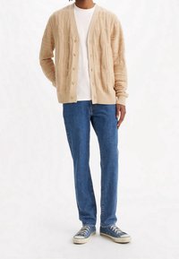 Beige stickad kofta med texturerad kabelmönster, med V-hals, knappframsida och ribbade ärmslut. Bärs över en vit skjorta och blå jeans.