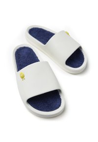 Sandalias deslizantes blancas con un forro de felpa azul marino, una correa ancha y un acento de un personaje sonriente amarillo en la correa. Material suave y liso.