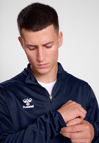 Sötétkék, cipzáros sportdzseki magas nyakkal, az előtérben fehér logóval a bal mellen, sima szövettextúrával. A modell igazít a hüvelyen.