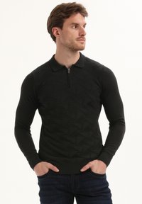 Vyras su rankomis kišenėse, vilkintis juodą tekstūruotą ilgomis rankovėmis polo megztinį ir tamsias džinsus, žiūri į dešinę.