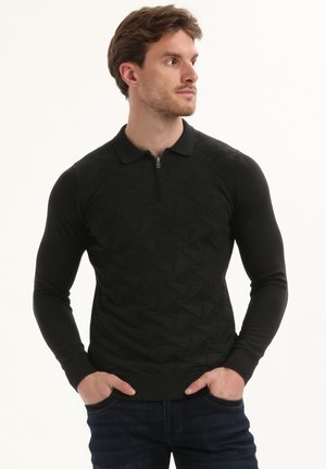 Uomo con le mani in tasca che indossa un maglione polo a maniche lunghe nero con texture e jeans scuri, che guarda verso destra.