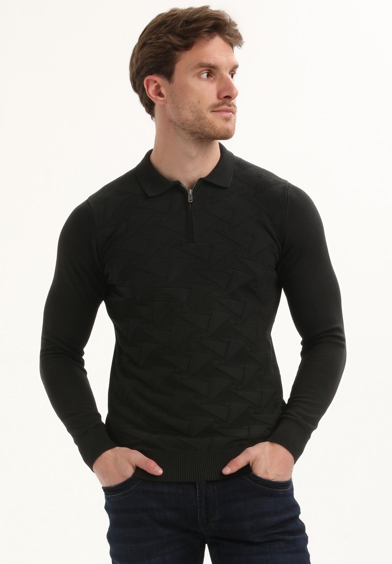 Vyras su rankomis kišenėse, vilkintis juodą tekstūruotą ilgomis rankovėmis polo megztinį ir tamsias džinsus, žiūri į dešinę.