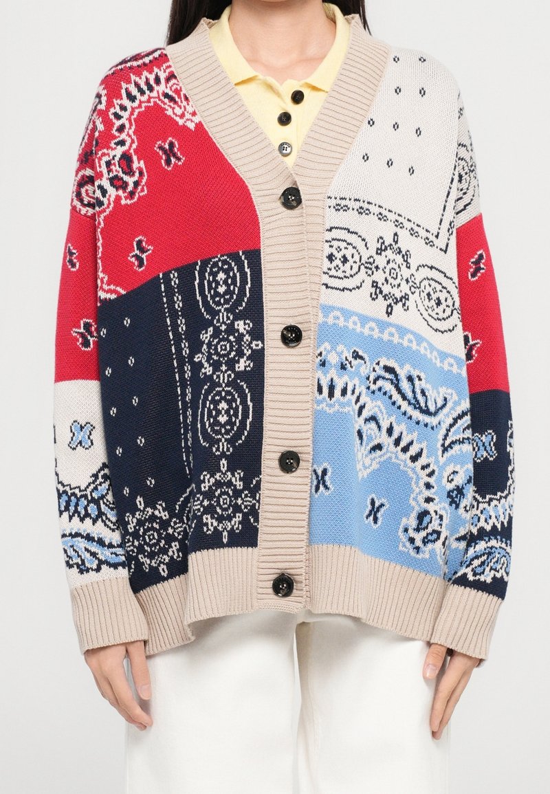 Femme portant un cardigan boutonné avec des motifs en patchwork rouges, marine, blancs et bleus, sur une chemise jaune à col.