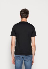 Musta puuvillahihaton t-paita, jossa on lyhyet hihat ja pyöreä pääntiukka, nähtynä takaa. Yhdistetty sinisiin farkkuihin, joissa on taskujen ompelut.