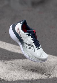 Άσπρο και μπλε ναυτικό αθλητικό παπούτσι τρεξίματος με κόκκινες λεπτομέρειες που αιωρείται πάνω από άσφαλτο με λευκές οδικές σημάνσεις.