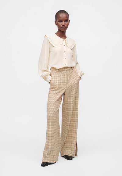 Femme portant un chemisier crème avec un col à volants et un pantalon large à carreaux beige, debout les mains dans les poches sur un fond uni.