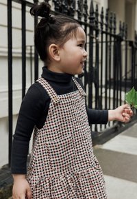 Bambino indossa un dolcevita nero sotto un abito senza maniche a quadri, con bottoni, orlo arricciato e una tasca frontale, tenendo una foglia.