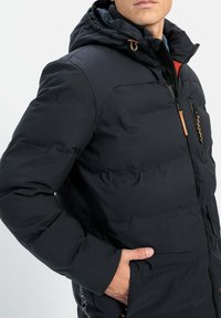Czarna pikowana kurtka z kapturem, z przodem na zamek błyskawiczny, zapinaną kieszenią i pomarańczowymi akcentami. Gładka tekstura i zdefiniowany krój.