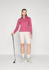 Kurzärmliges, rosa Oberteil mit Reißverschluss und lockerem Schnitt; beigefarbene Shorts bis zum Knie; hält einen Golfschläger; hellfarbene Sportschuhe.