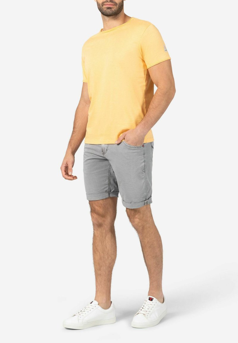 Gelbes T-Shirt mit kurzen Ärmeln, kombiniert mit grauen Shorts. Die Shorts haben umgeschlagene Säume, und das Outfit wird mit weißen Sneakers abgerundet.