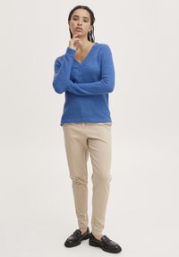 Zila V veida svītra, izgatavota no mīksta materiāla, apvienota ar bēša krāsas šauriem bikšu un melniem loafers. Modelis stāv ar vienu roku uz zoda.