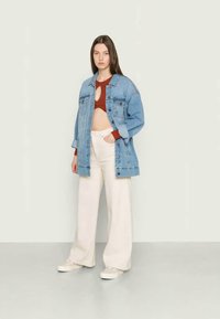 Casaco de ganga em azul claro com dois bolsos frontais, usado sobre um top castanho curto, combinado com calças largas em tom creme e ténis.