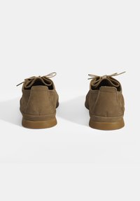 Chaussures en suède marron avec des orteils arrondis, des œillets proéminents et des lacets en nœud. Des panneaux latéraux texturés et des semelles en caoutchouc complètent le design.