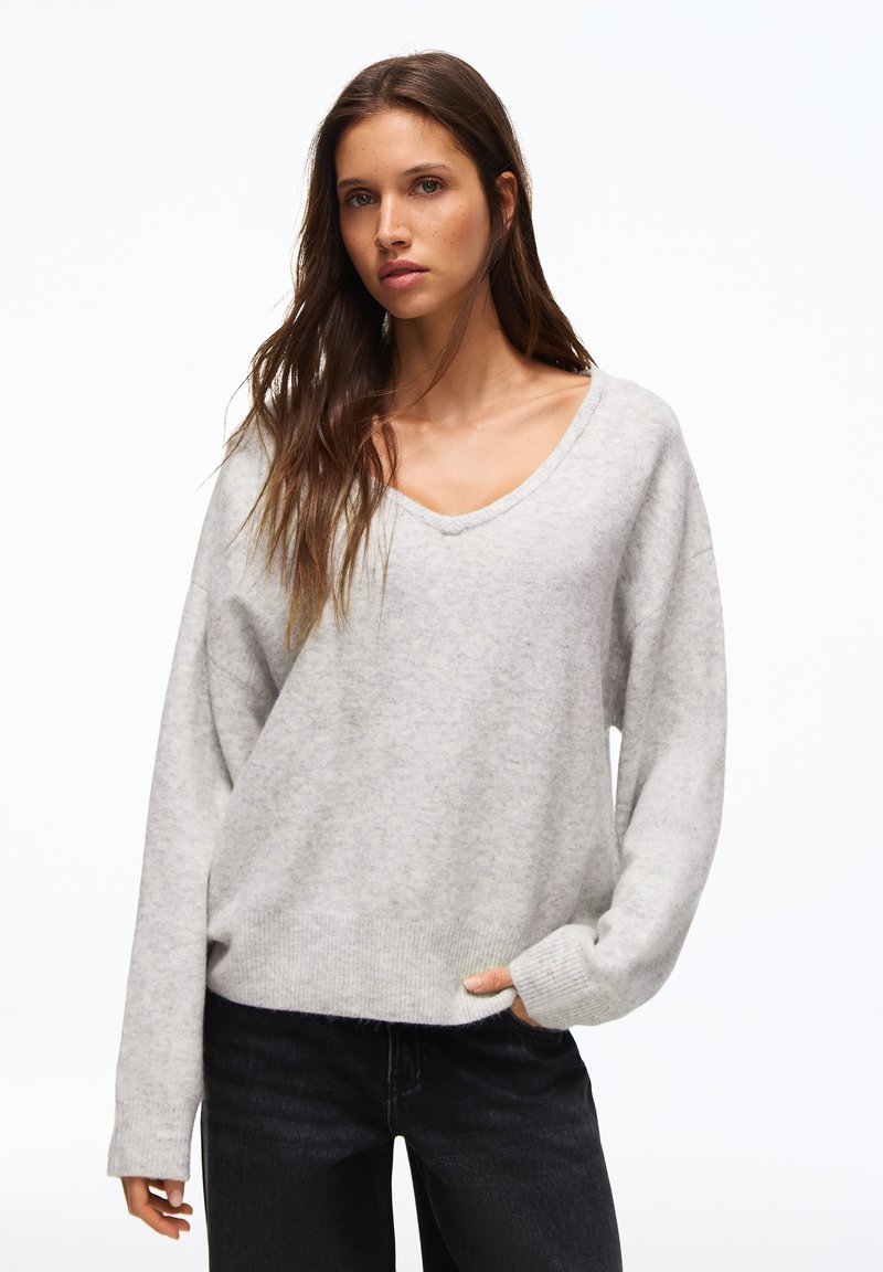 Jeune femme aux longs cheveux bruns portant un pull gris clair à col en V et un pantalon noir sur un fond blanc uni.