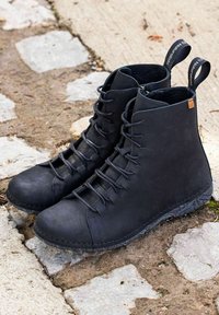 Botines negros con cordones y tiradores etiquetados "El Naturalista", colocados sobre una superficie exterior de piedra y tierra.