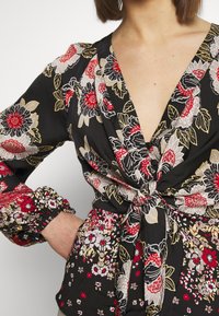 Black floral blouse featuring red, white, and gold patterns, deep V-neck, and tie waist detail, with long, puffed sleeves and elastic cuffs.