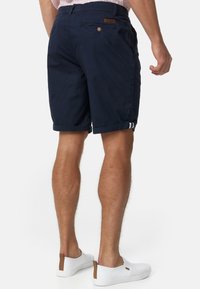 Shorts en coton bleu marine avec des revers roulés, deux poches arrière et une étiquette en cuir. Les accessoires comprennent des chaussures blanches à enfiler avec un accent beige.