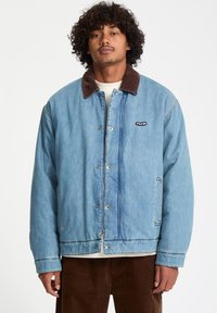 Lichtblauwe denimjas met een bruine corduroy kraag, vooraan drukknoopsluiting en zijzakken, met een klein zwart logo-opdruk op de borst.